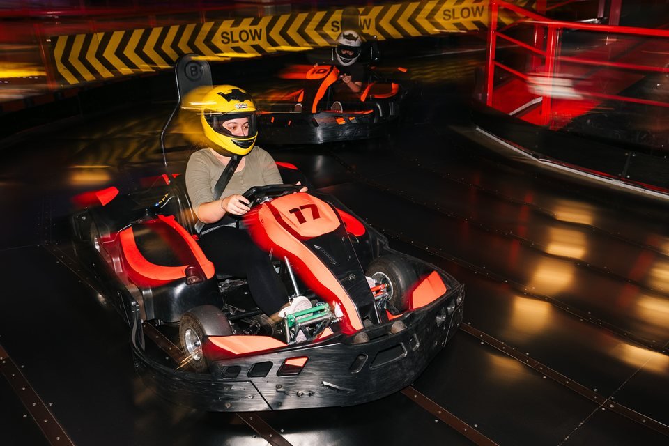 Two karts on the lower level at Grand Pier Go-Karts, passing the black-and-yellow SLOW chevrons on the barrier
