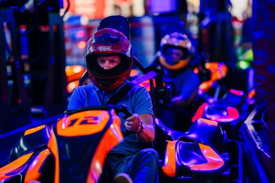 Two drivers in helmets racing orange karts under the purple and blue lighting at Grand Pier Go-Karts