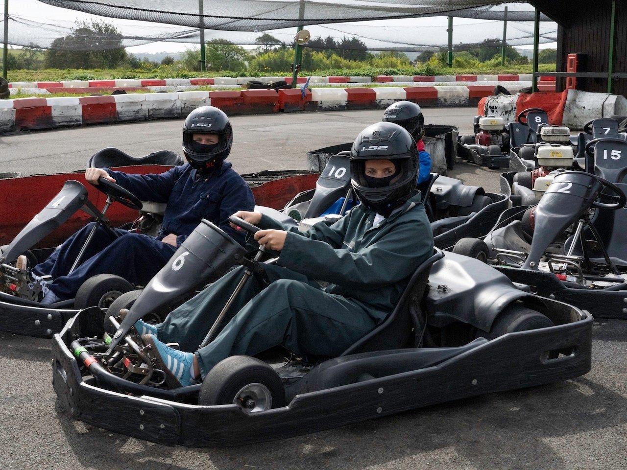 Drivers in overalls and helmets waiting in the karts beside the pit shelter at Heatherton World of Activities, with the outdoor circuit behind