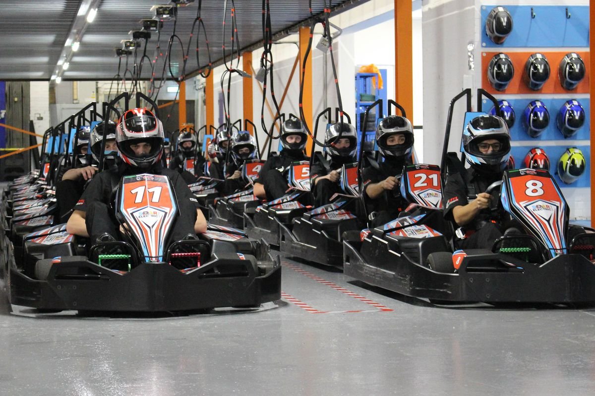 A full grid of drivers lined up in the pit lane at SOAR Electric Karting, with the helmet rack on the wall behind