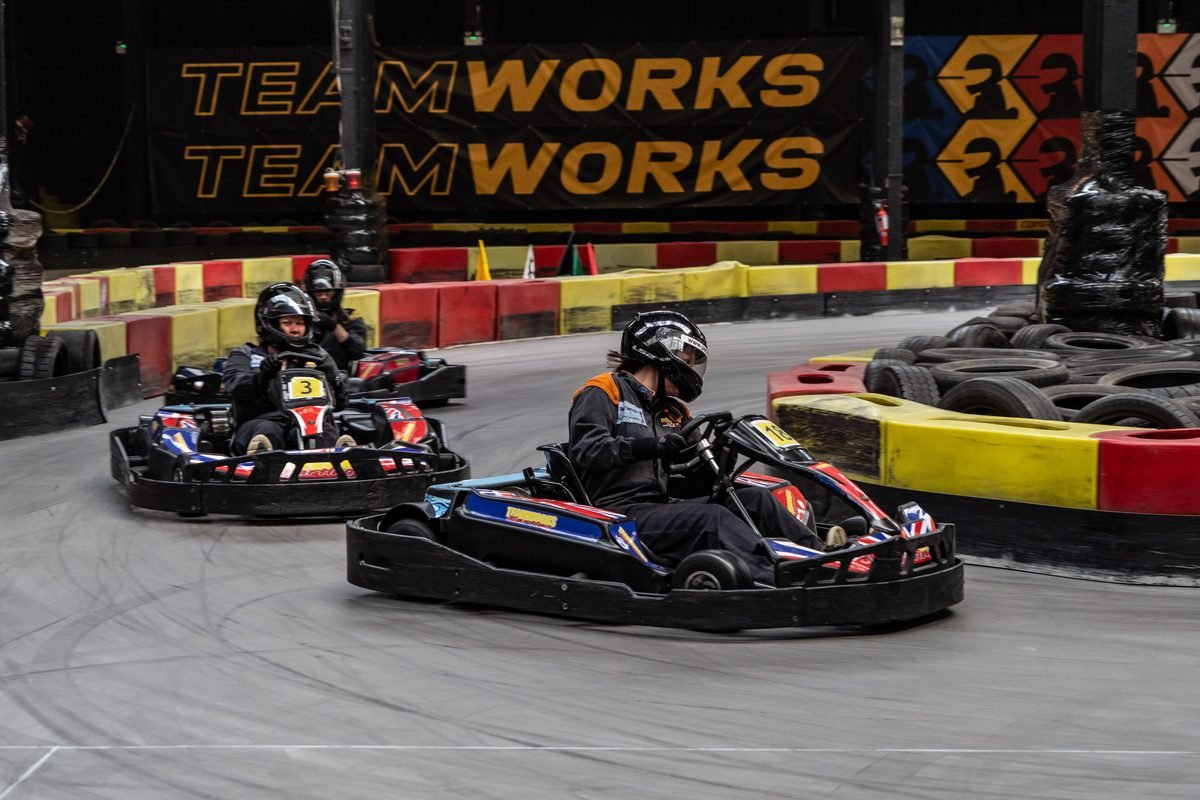 Two karts racing side by side on the indoor circuit at Teamworks Karting Birmingham City, with Teamworks branding on the barriers behind